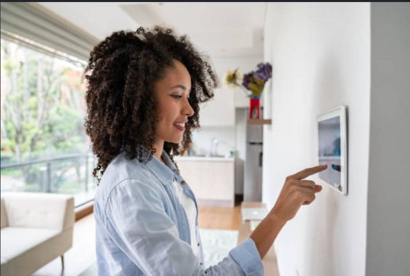 A woman adjusting her home from a wall-mounted touch panel in a bright open-plan living room